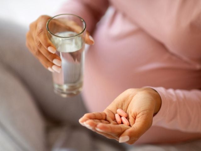 Pregnant person holding a glass of water and pink pills in one hand.